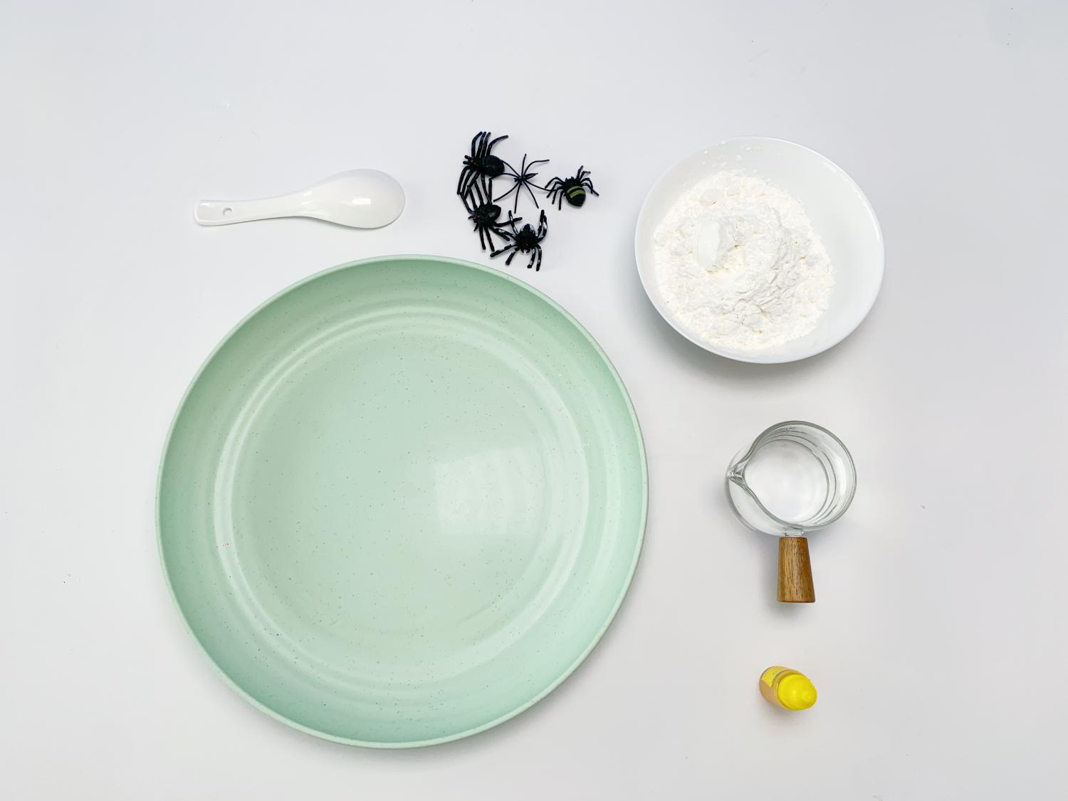 Several items are arranged on a white surface, clearly prepared for a Halloween-themed craft activity. A pale green, shallow dish is centrally positioned, alongside a small white bowl containing a white powder (likely cornflour), a small glass jug with clear liquid (likely water), a white spoon, a small yellow container (probably food colouring), and a group of small, black plastic spiders. The arrangement suggests the preparation for making a Halloween-themed sensory activity, possibly Oobleck, given the cornflour and water. The overall presentation is neat and organised, ready for the craft to begin.