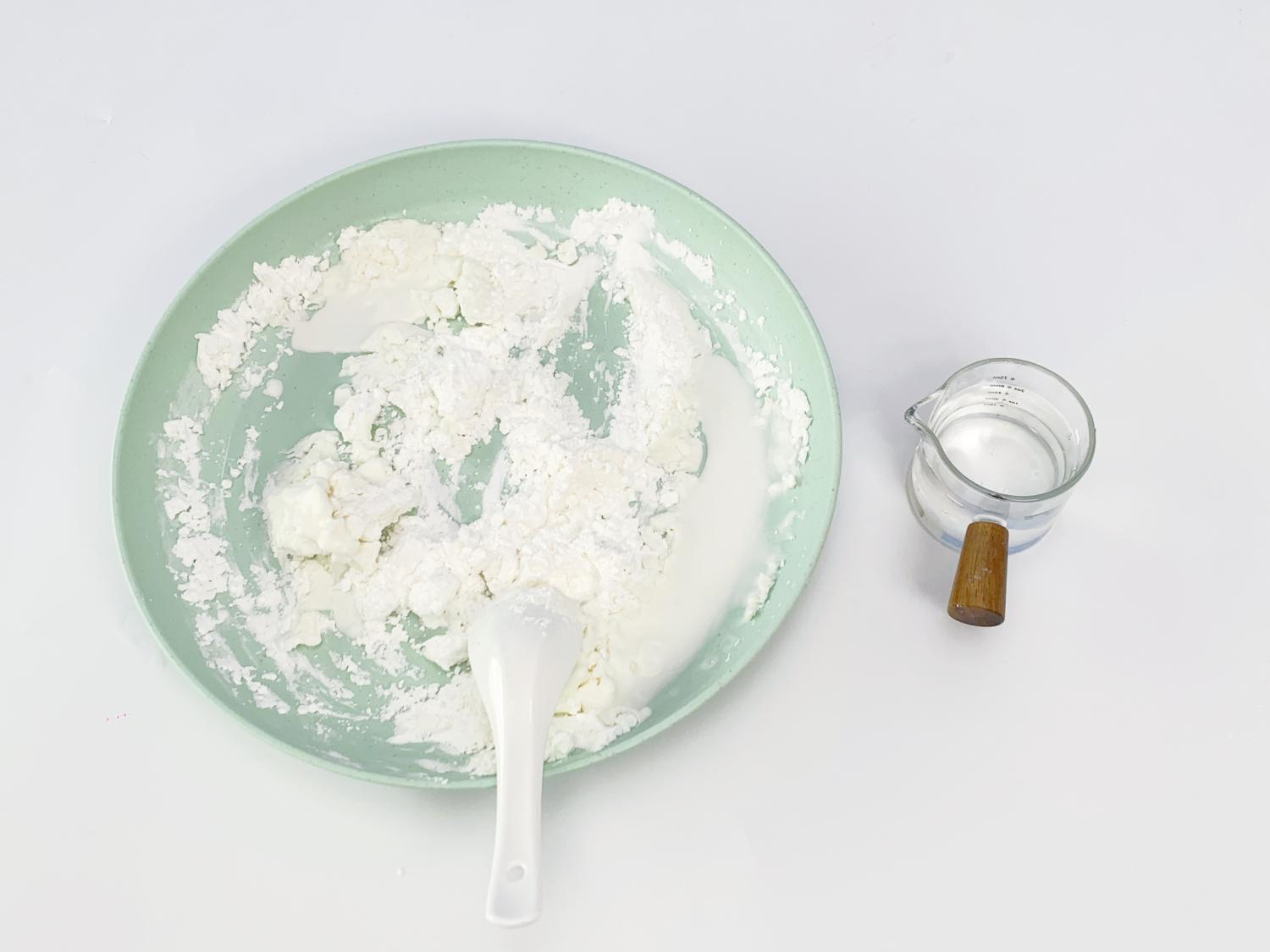 A pale green dish holds a mixture of white powder and a creamy white liquid, likely cornflour and water in the process of being combined to make Oobleck. A small white spoon rests within the mixture. A small glass jug containing more clear liquid sits to the right of the dish. The background is a plain white surface. The image shows a partially completed stage in making Oobleck, a non-Newtonian fluid, highlighting the initial mixing process.