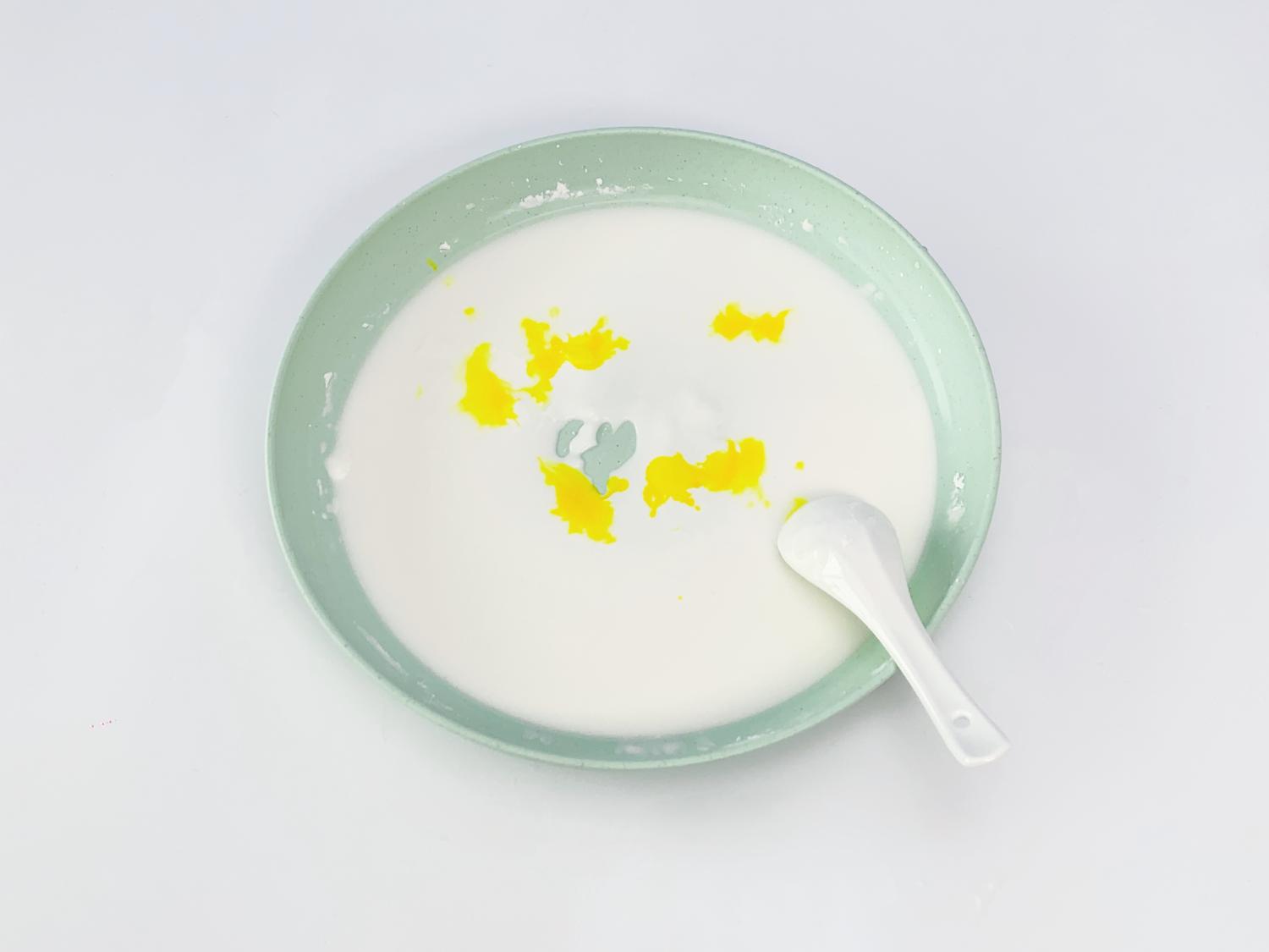 A pale green dish contains a mixture of white Oobleck, with several splashes of yellow food colouring unevenly distributed throughout. A white spoon rests within the mixture, suggesting it has just been used for stirring. The background is a plain white surface. The image shows a step in the process of making coloured Oobleck, highlighting the addition and mixing of the food colouring. The overall impression is of a simple, hands-on science or craft activity. 