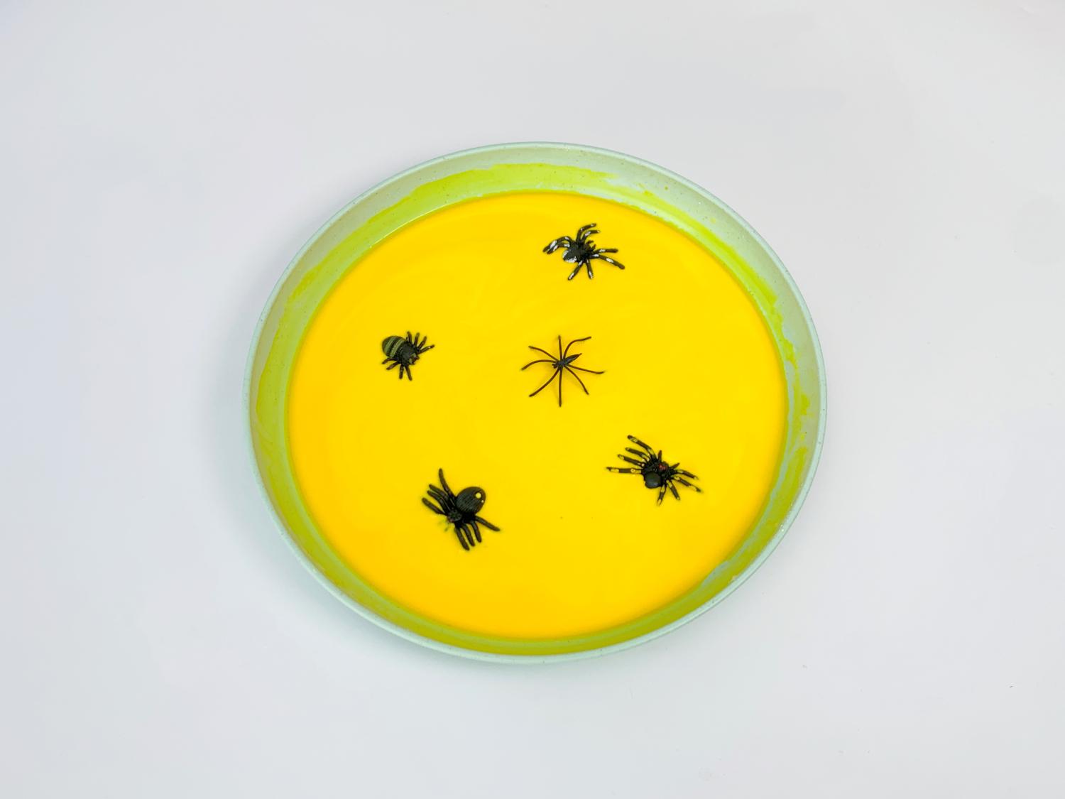 A pale green shallow dish contains a vibrant yellow, viscous substance, possibly Oobleck. Several small, black plastic spiders are partially submerged within the mixture. The background is a plain white surface. The image presents a completed Halloween-themed sensory activity, highlighting the contrast between the bright yellow Oobleck and the dark spiders. The overall impression is one of a fun and slightly spooky tactile experience, suitable for children.