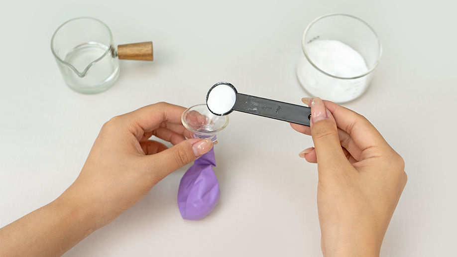 Adding the magic ingredient! A pair of hands carefully pours bicarbonate of soda from a measuring spoon into a small funnel held over a deflated purple balloon. It's all systems go for some spiky science fun!