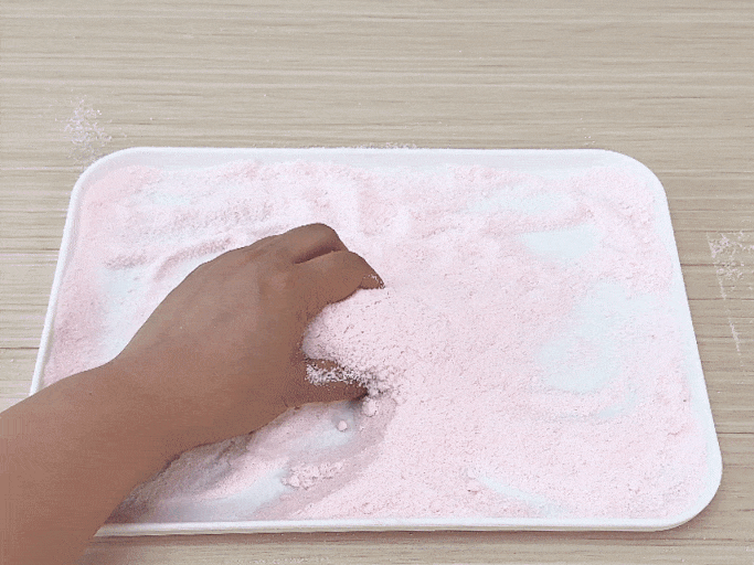 A hand sprinkles light pink powder onto a white tray. The powder accumulates in a small pile. Then, a hand uses a butter knife to cut a round, compacted disc of the same pink powder, which is nestled within the loose powder on the tray. The disc is sliced into several sections, resembling the cutting of a cake or pie. The video demonstrates a simple activity involving powdered material, possibly bath bomb mixture or a similar substance.
