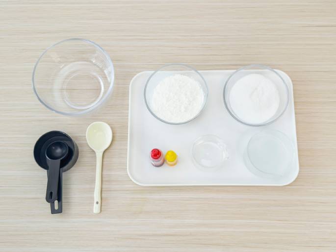 An assortment of ingredients and tools are laid out on a light woodgrain table, suggesting preparation for baking or a science experiment. A large clear glass bowl sits to the left. In the center, a white rectangular tray holds several smaller clear glass bowls containing what appears to be flour, sugar, and water. Two small bottles of red and yellow food coloring are also on the tray. To the left of the tray are a set of black measuring spoons and a cream-colored mixing spoon. The overhead view presents all items clearly, giving the impression of organized preparation.