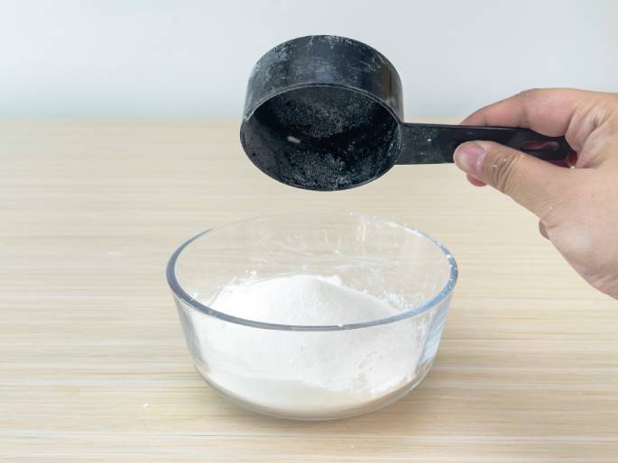 A hand holds a black measuring cup containing white powder, likely flour or sugar, over a clear glass bowl on a light woodgrain surface. The bowl already contains some of the white powder. The image suggests baking or cooking preparation. A plain off-white background keeps the focus on the action.