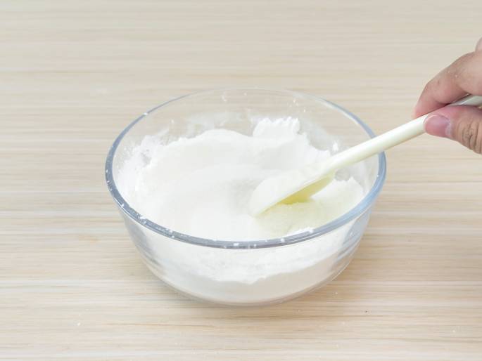 A clear glass bowl containing a white powder, likely flour or cornstarch, is being mixed with a small amount of liquid using a cream-colored spatula. A hand holds the spatula, stirring the mixture. The bowl sits on a light woodgrain table, and a plain off-white background keeps the focus on the process. The image suggests baking, cooking, or a science experiment involving mixing ingredients.