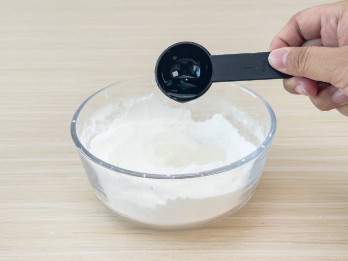 A hand pours a clear liquid from a small, black measuring spoon into a clear glass bowl containing a white powder, likely flour or cornstarch. The bowl sits on a light woodgrain table. The scene suggests a step in a recipe or a science experiment where ingredients are being combined. A plain, off-white background keeps the focus on the action.
