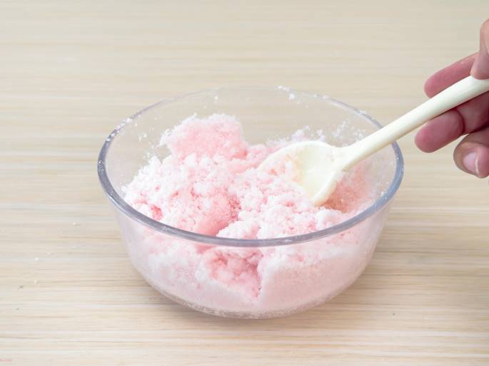 A clear glass bowl filled with a crumbly, light pink mixture sits on a light woodgrain table. A cream-colored spoon is being used to mix the contents, and a hand is visible holding the spoon. The image suggests a baking or cooking process, perhaps involving dyed sugar or a similar ingredient. A plain, off-white background keeps the focus on the bowl and its contents.