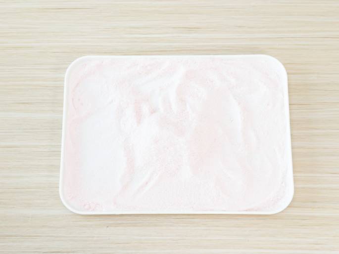 A white rectangular tray contains a layer of predominantly white powder with streaks of light pink mixed in. The tray rests on a light woodgrain surface. The image suggests a baking or cooking process, perhaps involving dyed sugar, flour, or a similar ingredient being prepared or spread out.