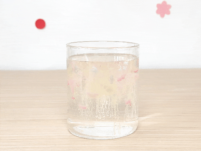 A clear glass sits on a light wooden surface, filled with a clear liquid. Pastel-colored inks or dyes, in shades of pink, yellow, and light blue, are slowly diffusing downwards, creating wispy, colorful trails. The background is a light-colored wall with a red dot and a pink flower shape, likely decorations. The video showcases the visual effect of the colors mixing with the liquid.