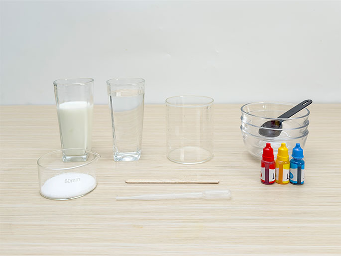 An assortment of ingredients and tools are arranged on a light woodgrain table, likely for a science experiment or cooking activity. From left to right: a glass filled with milk, a glass of water, an empty cylindrical glass container, a stack of clear glass bowls with a small black measuring spoon inside the top bowl, three small bottles of food coloring (red, yellow, blue), a wooden craft stick, and a clear plastic pipette or dropper. A small glass beaker containing a white powder, possibly sugar or salt, is positioned in front of the milk and water glasses. The beaker has 80mm marked on its side.