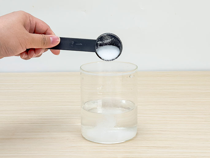 A hand holds a black measuring spoon filled with white granules, likely salt or sugar, over a clear glass containing water. The granules are being poured into the water, with some already visible at the bottom of the glass. The glass sits on a light woodgrain surface. This image likely depicts the process of dissolving a substance in water.