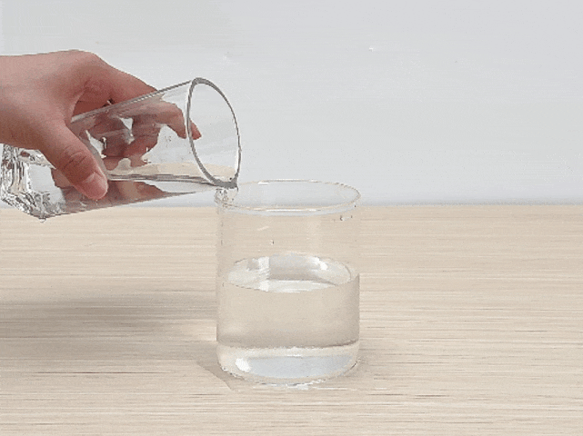 A hand pours water from a clear drinking glass into a shorter, cylindrical clear glass on a light woodgrain surface. The cylindrical glass is already partially filled with water.