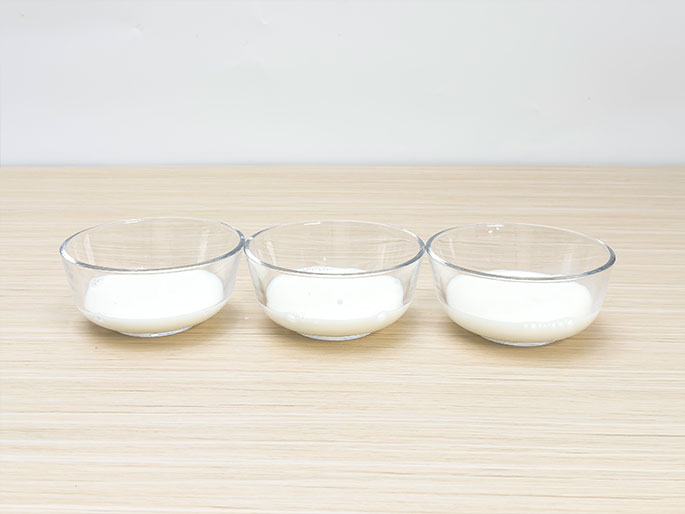 Three small, clear glass bowls containing milk are arranged in a row on a light woodgrain table. Each bowl holds a similar amount of milk. They appear ready for an experiment or recipe.