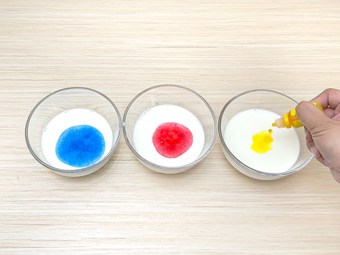 Three clear glass bowls containing milk sit on a light woodgrain table. Drops of blue, red, and yellow food coloring have been added to each bowl, creating distinct circles of color on the surface of the milk. A hand is shown in the process of adding yellow food coloring to the third bowl. This setup likely demonstrates a surface tension experiment or a colorful activity involving milk and food coloring.