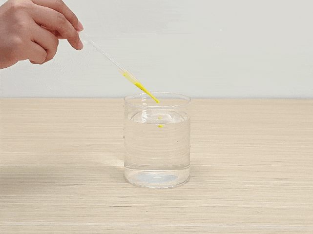 A hand holds a clear plastic pipette containing yellow liquid, likely food coloring, over a clear glass partially filled with water. Droplets of the yellow liquid are being dispensed from the pipette into the water. The glass sits on a light woodgrain surface. This image likely depicts an experiment or demonstration involving color mixing or diffusion.