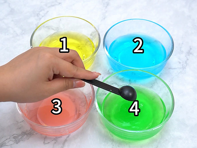 Two hands are shown pouring liquid from a small, clear measuring cup onto a piece of clear plastic sheeting. The sheeting is held taut between the hands, and the liquid is being poured onto its center. The background is a light wood-grain surface.