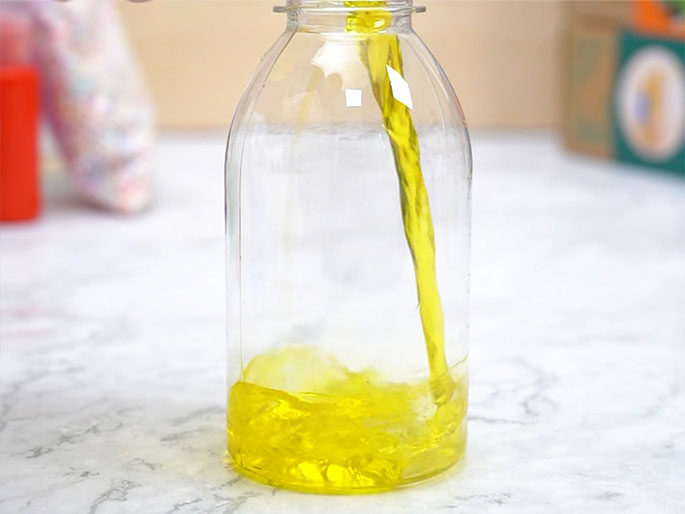 Two hands hold a small, clear plastic pouch filled with a clear liquid. The pouch is tied off at the top with a knot, which one hand is pinching and twisting. The background is a light wood-grain surface.