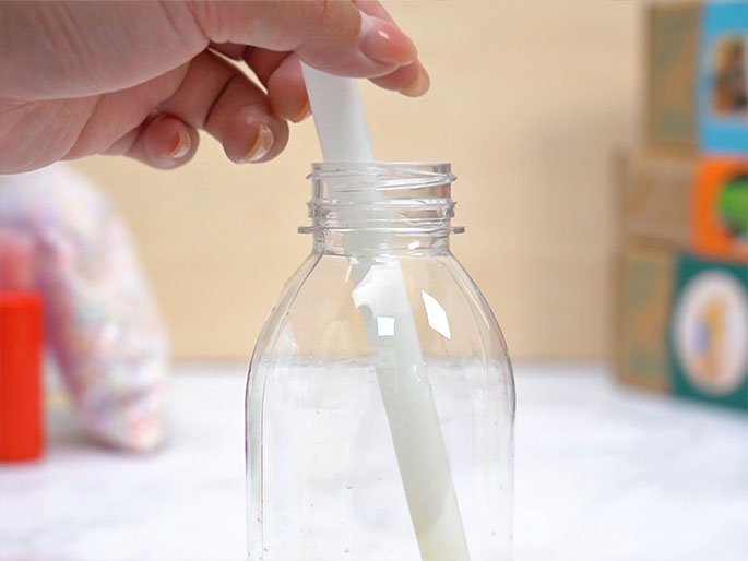 A hand holds a small, clear, water-filled plastic pouch tied at the top. A pair of scissors is positioned to trim the excess plastic just below the knot. The background is a light wood-grain surface.