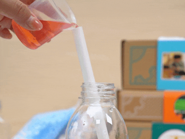 A hand pours a vibrant reddish-orange liquid from a clear measuring cup into a clear plastic bottle. A white, straw-like object is positioned inside the bottle's opening, guiding the liquid's flow. In the background, several cardboard boxes are stacked, each featuring colorful labels with images that suggest they contain science experiment materials. A piece of light blue cloth is partially visible beneath the bottle. The video showcases the process of preparing a science experiment, likely involving colored liquids.