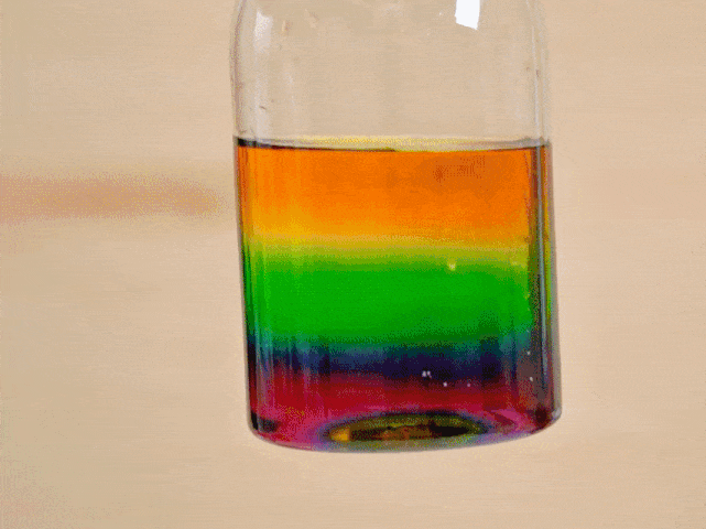 A clear glass bottle is filled with liquids layered to resemble a rainbow. The distinct horizontal bands of color, from top to bottom, are red, orange, yellow, green, blue, indigo, and violet. The bottle is gently rotated, causing the colorful liquids to swirl and mix slightly at the interfaces between the layers. This visually appealing demonstration likely illustrates the concept of liquid density or layering liquids based on their different properties.