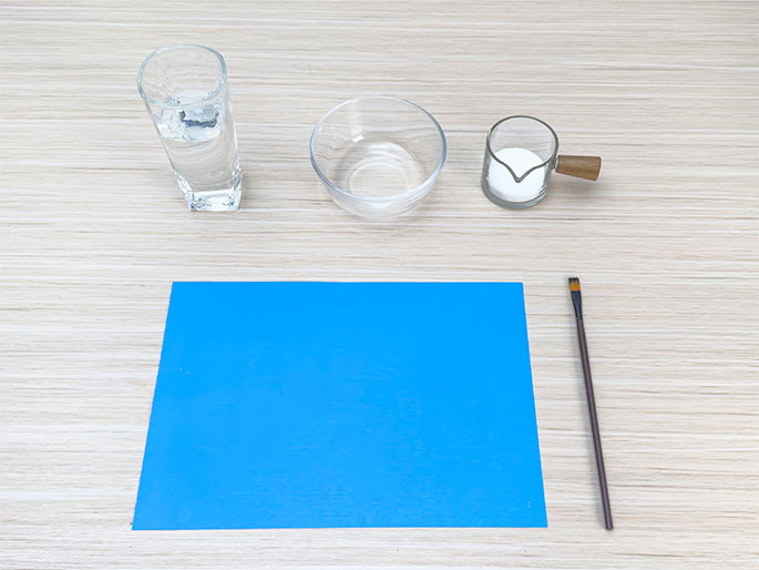Laid out on a light wood-grain table are several items, likely for a science experiment or craft project. A clear glass filled with water sits at the left, followed by an empty clear bowl. Next to the bowl is a small, clear creamer-shaped container with a wooden handle, containing a white substance, possibly sugar or salt. Below these items is a rectangular piece of bright blue material, possibly paper or foam. A thin paintbrush with a dark handle and light bristles lies to the right of the blue material. The arrangement suggests preparation for an activity involving mixing or applying substances.
