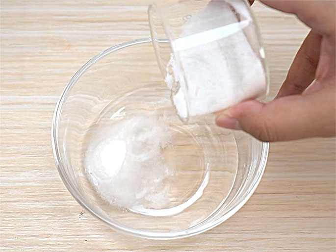A hand pours a white powder, likely sugar or salt, from a small, clear, creamer-shaped container with a wooden handle into a clear glass bowl. The bowl sits on a light wood-grain surface. Some of the powder has already been poured into the bowl, and the image captures the process of adding more. This close-up shot suggests a step in a recipe, experiment, or craft activity.