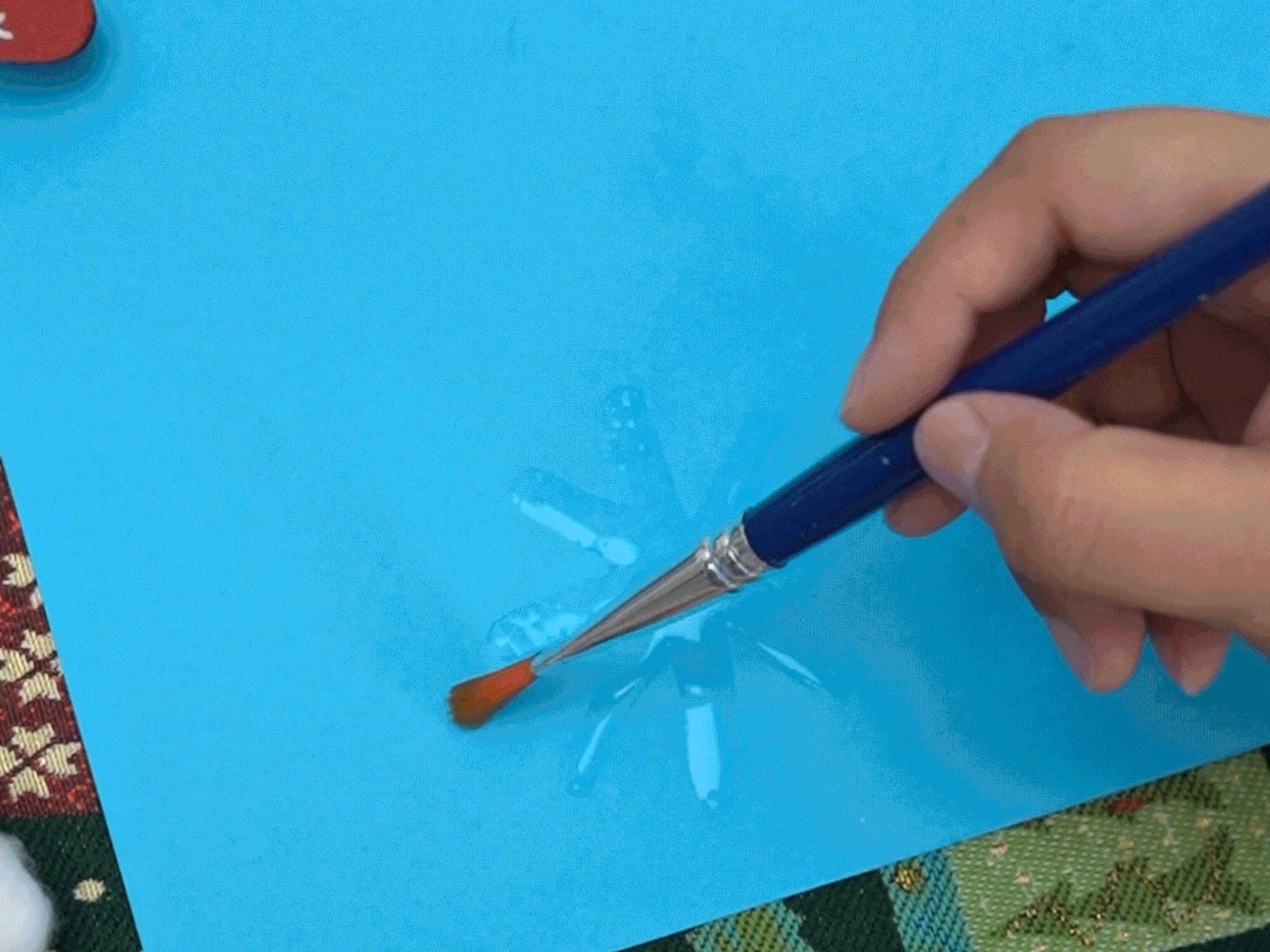 A hand guides a paintbrush with an orange tip across a light blue surface. The brush seems to be applying a clear or slightly glossy substance onto a snowflake design, making it appear wet and shiny. The background features glimpses of patterned fabrics in red, brown, and green, along with a touch of white, possibly cotton.
