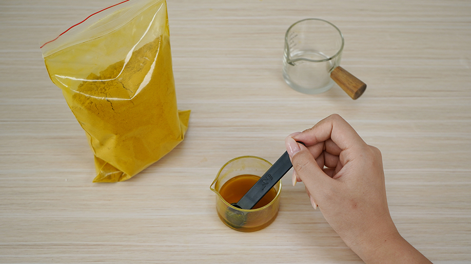 A hand carefully adds a spoonful of turmeric to a small glass of water, creating the vibrant base for our invisible ink. It's all rather mysterious, innit?