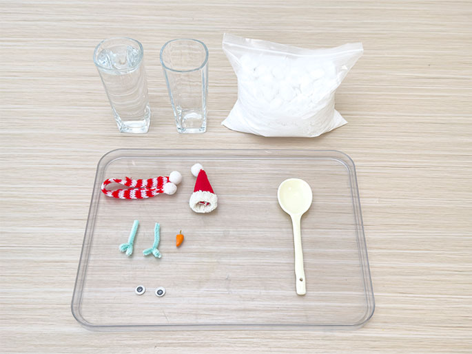 Arranged on a light wood-grain surface are materials for a snowman-making activity. At the top, two clear glasses, one filled with water and the other empty, sit beside a clear plastic bag containing a white substance, likely fake snow. Below, on a clear rectangular tray, are various snowman components: a miniature red and white striped scarf with white pom-poms, a small red Santa hat, two light green pipe cleaner arm pieces, a small orange carrot nose, two black button eyes, and a cream-colored spoon. This image depicts the ingredients and accessories ready for assembling a miniature snowman.
