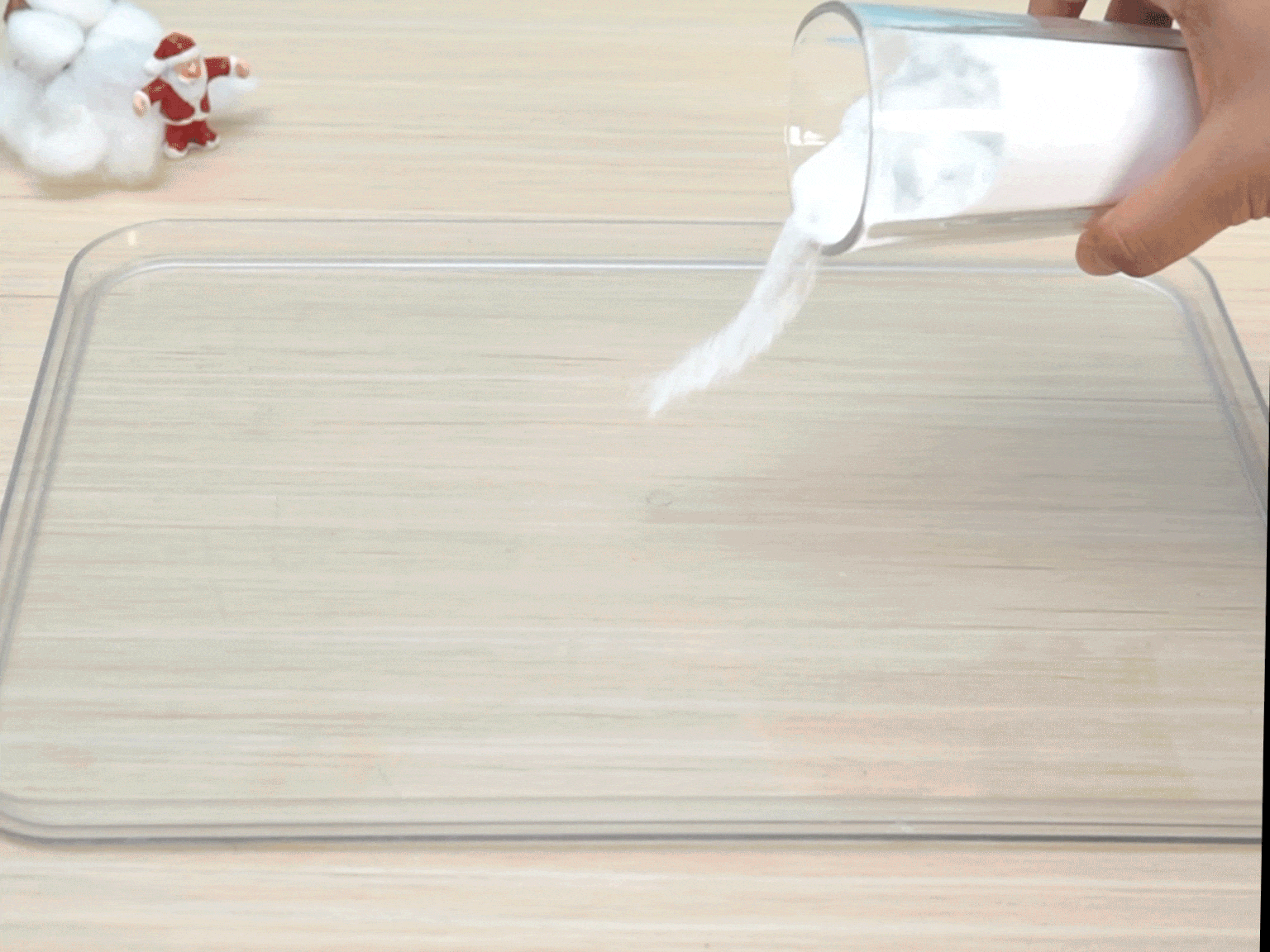 A hand pours white powder from a clear glass onto a clear plastic tray. The powder flows smoothly, creating a small pile on the tray's surface. The tray rests on a light wooden table. A small Santa Claus figurine and some white cotton are visible in the background, slightly out of focus.