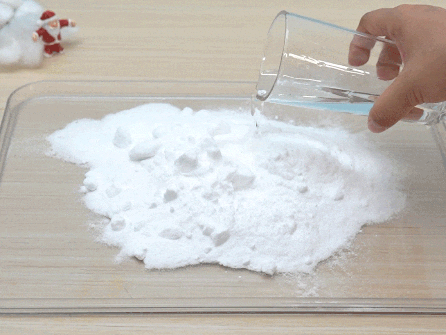 A hand pours water from a clear glass onto a pile of white powder on a clear plastic tray. The water mixes with the powder, likely causing a reaction or change in consistency. The tray sits on a light wooden table. A small Santa Claus figurine and some white cotton are visible in the background, slightly blurred.