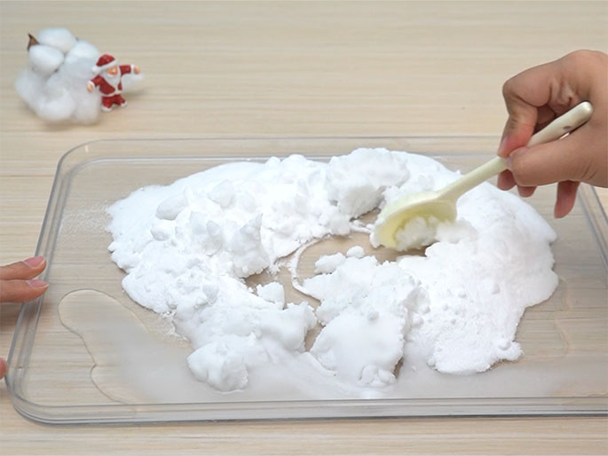 On a clear rectangular tray placed on a light wood-grain table, a hand uses a cream-colored spoon to mix a white powdery substance, likely fake snow, with a clear liquid. A small pile of fluffy white cotton and a miniature Santa Claus figure are visible in the background, out of focus, suggesting this is part of a Christmas-themed craft activity. The mixture on the tray is being combined to likely create a moldable snow-like material. A small puddle of the clear liquid is visible on the left side of the tray.