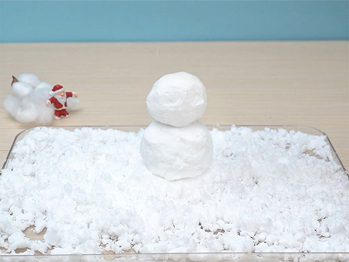 A small snowman made of two white balls, one stacked on top of the other, sits on a bed of white artificial snow spread across a clear rectangular tray. The background, a blend of light wood and a pale blue wall, is slightly out of focus. A small pile of white cotton and a miniature Santa Claus figurine are also visible, blurred in the background to the left. The image captures a simple, festive scene, likely a craft project or a decorative element.