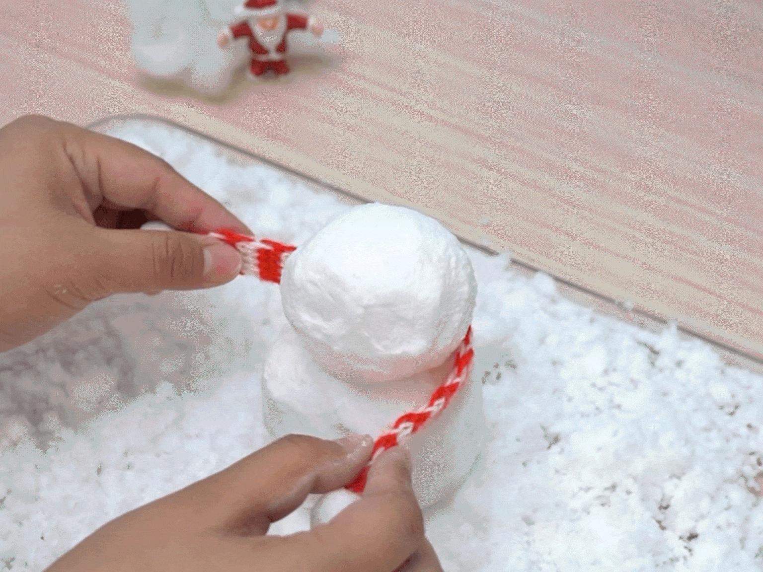 Hands are shown placing a red and white striped scarf around a small snowman made of white material, possibly snow or dough. The snowman is positioned on a bed of white, powdery material, resembling snow, on a clear tray. The tray rests on a light wooden surface. A small Santa Claus figurine is visible in the blurred background.