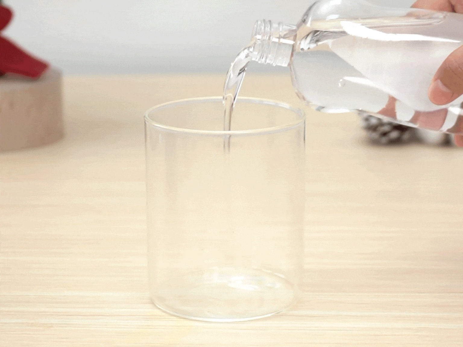A clear plastic bottle is being tilted, pouring a clear liquid into a clear glass tumbler. The glass sits on a light-colored wooden table. A blurred red object and a pine cone are visible in the background, slightly out of focus.