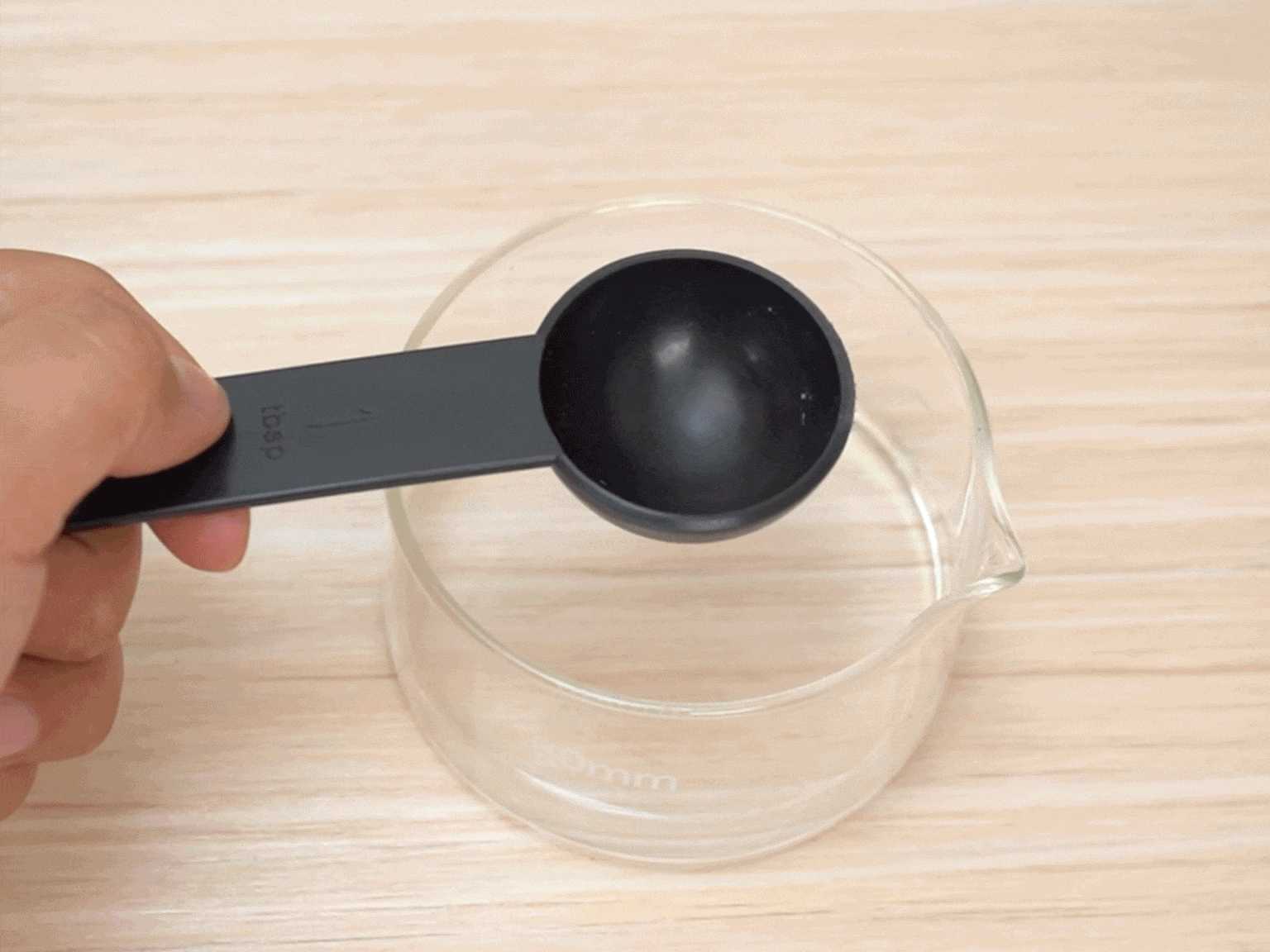 A hand holds a black plastic measuring spoon, labeled 1 tbsp, over a clear glass container with a small amount of clear liquid inside. The background is a light woodgrain surface. The spoon appears to be about to add the measured ingredient to the container.