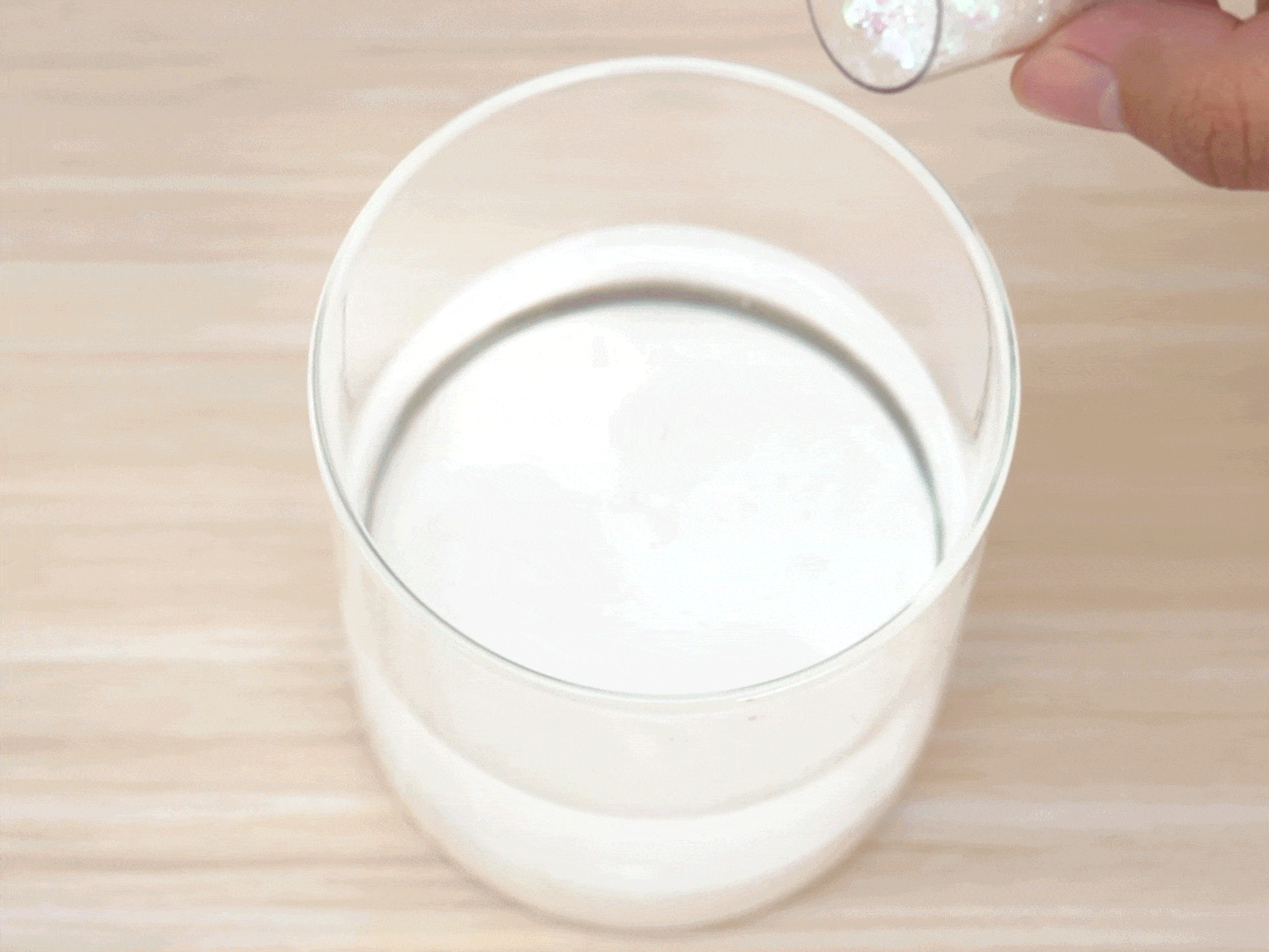 A hand holds a small, clear container of iridescent glitter and pours it into a clear glass tumbler. The glass contains a milky white liquid, likely a mixture of glue and water. The glass sits on a light woodgrain table. This image likely depicts a step in a crafting project.