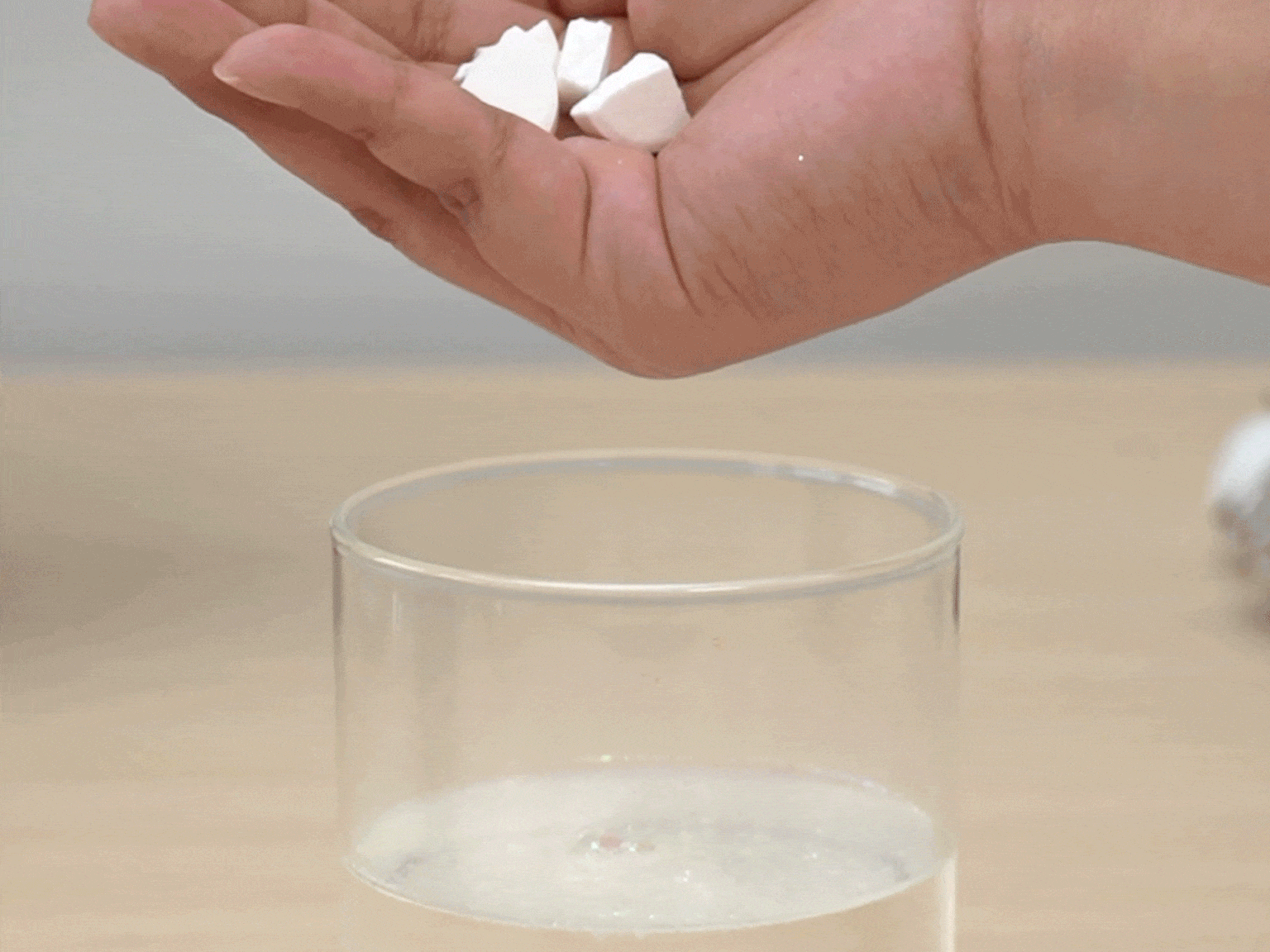 A hand holds several small, white, irregularly shaped pieces of a substance, possibly borax, above a clear glass tumbler containing a clear liquid. The pieces are being dropped into the glass. The glass sits on a light wood surface. A blurred white object is visible in the background, out of focus. This image likely depicts a step in a science experiment or DIY project.