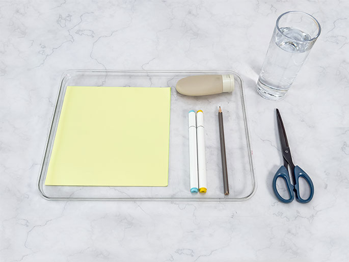 Arranged on a clear plastic tray are several art supplies: a pale yellow sheet of paper, two white markers with light blue and yellow caps, a pencil, and a small beige squeeze bottle. A clear glass of water and a pair of blue-handled scissors sit beside the tray on a marbled surface.