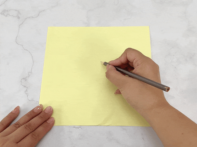A hand holding a gray pencil begins to draw a spiral on a square sheet of pale yellow paper. The paper rests on a marbled surface. The hand starts the spiral from the center of the paper and continues to draw outwards in a clockwise direction. The video focuses on the process of drawing this spiral shape.
