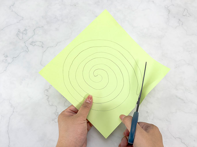 Two hands hold a square of pale green paper with a pencil-drawn spiral on it. One hand holds the paper steady while the other hand uses scissors to cut along the spiral line. The background is a marbled surface.