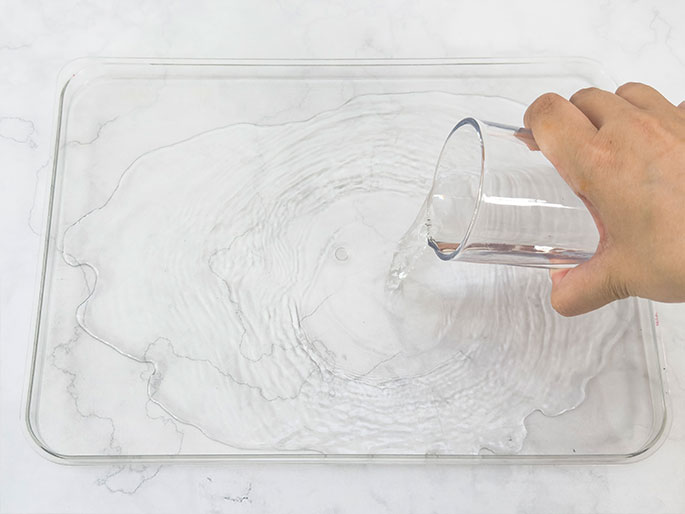 A hand pours water from a clear glass into a clear rectangular tray. The water creates ripples and spreads across the tray's surface. The tray sits on a marbled background.