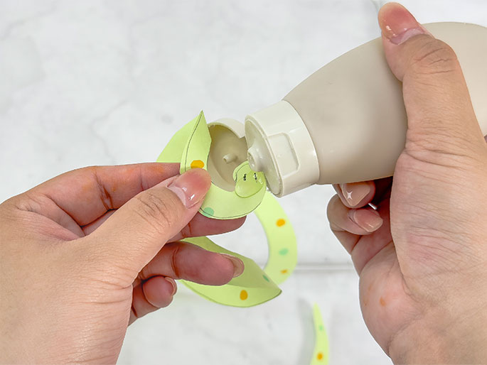 Two hands work together to apply glue from a beige squeeze bottle to the end of a light green paper spiral decorated with orange and green dots. The spiral is partially unfurled, and the end being glued has a simple face drawn on it, resembling a snake.