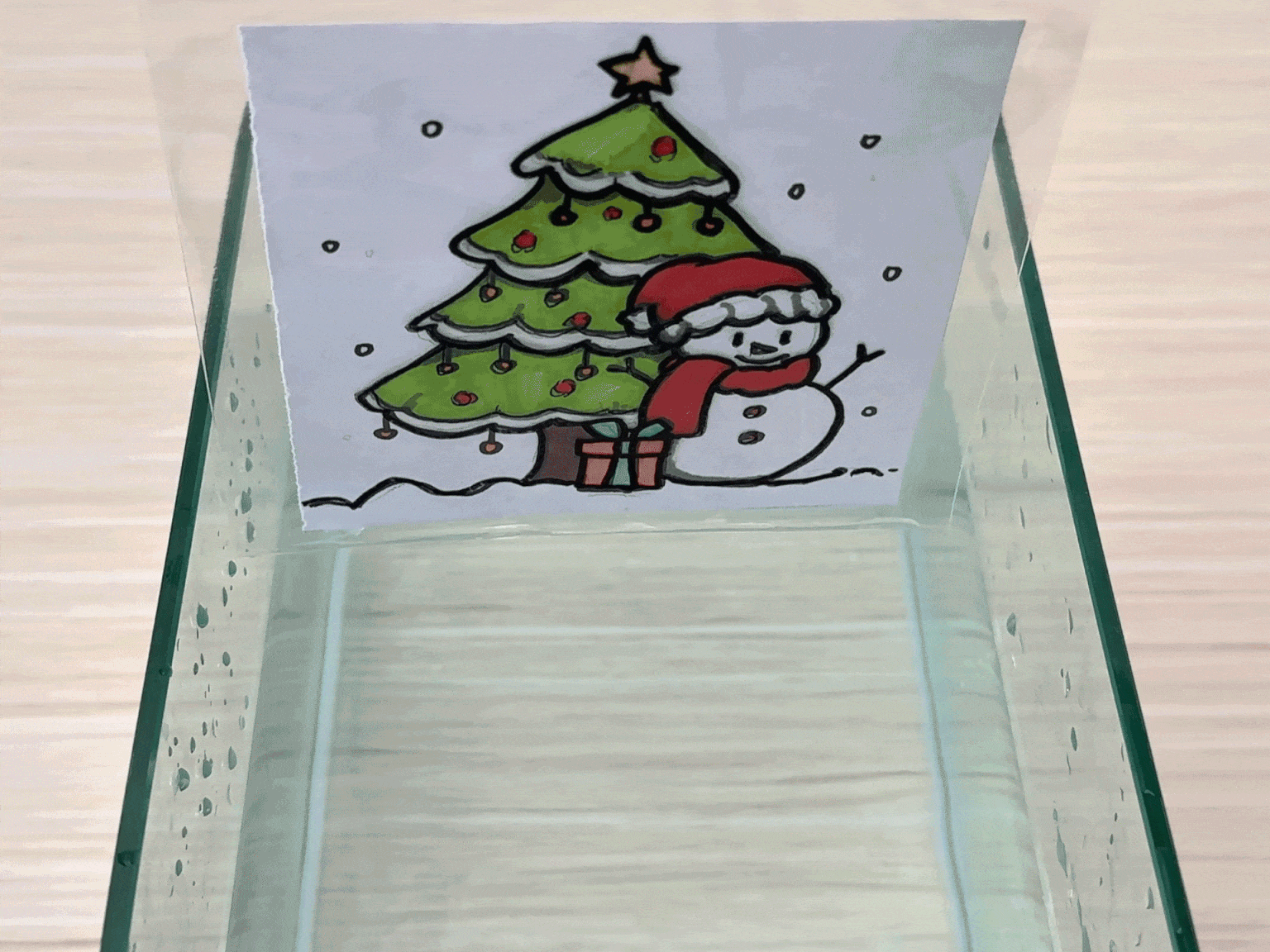 A hand holds a small, clear plastic bag containing a drawing of a Christmas scene. The drawing depicts a decorated Christmas tree and a snowman wearing a Santa hat in a snowy setting. The bag is partially submerged in a clear container filled with water. The hand moves the bag slightly, causing the image inside to appear to move and distort due to the refraction of light through the water. The video demonstrates a simple visual experiment or activity involving water and a plastic bag.