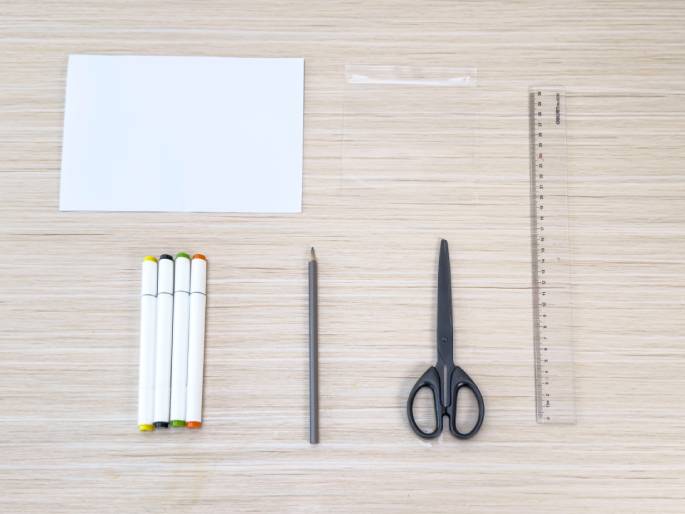 A flat lay arrangement of craft supplies on a light woodgrain table. The supplies include a blank sheet of white paper, a small clear plastic bag, a clear plastic ruler, a gray pencil, a pair of black scissors, and four white markers with colored caps (black, green, yellow, and orange). The items are neatly arranged, suggesting preparation for a craft project.