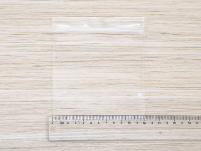 A small, empty, clear plastic bag with a zipper seal lies on a light woodgrain table. A clear plastic ruler is positioned below the bag, likely to indicate its size.