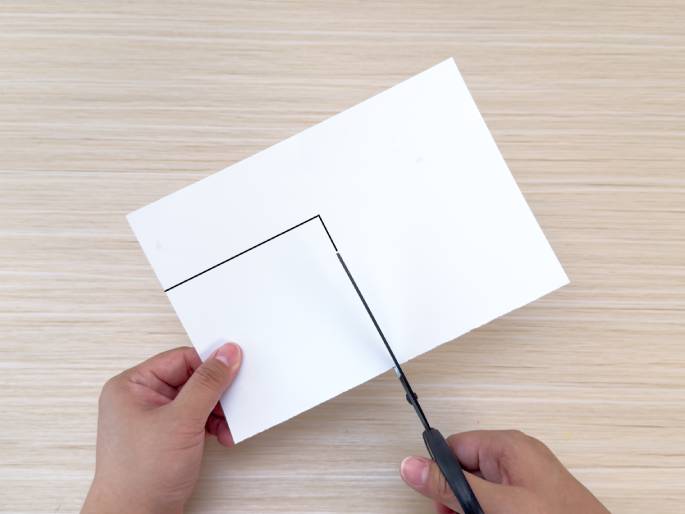 Two hands are shown cutting a piece of white paper with black scissors. The paper rests on a light woodgrain table and has a black line drawn on it, guiding the cut. The hands are holding the paper and scissors in position to cut along the line. This image likely depicts a step in a craft or DIY project.
