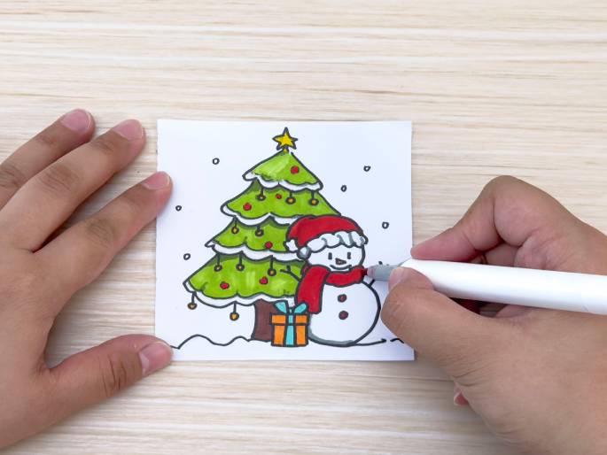 A hand holds a white marker and colors a drawing of a Christmas scene on a small white piece of paper. The drawing depicts a decorated green Christmas tree with a yellow star on top, a snowman wearing a red Santa hat and scarf, a wrapped present, and falling snow. The paper rests on a light woodgrain table. Another hand is visible, holding the paper steady.