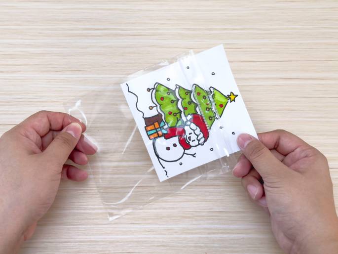 Two hands are placing a drawing into a small, clear plastic bag with a zipper seal. The drawing depicts a Christmas scene with a decorated tree, a snowman wearing a Santa hat and scarf, a present, and falling snow. The bag is being sealed to protect the drawing. The background is a light woodgrain surface.