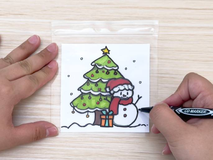 A hand holds a black marker and adds details to a drawing enclosed in a clear plastic bag. The drawing, visible through the bag, features a Christmas tree, a snowman with a Santa hat and scarf, a present, and falling snow. Another hand holds the bag steady. The bag and drawing rest on a light woodgrain table. This image seems to depict creating a simple, protected Christmas decoration.